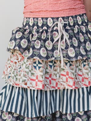 A close-up detail shot of a tiered ruffle skirt or pant in a playful mix of prints — floral medallion, patchwork geometric, and vertical stripe — layered together with a drawstring waist tie, paired with a coral textured top.