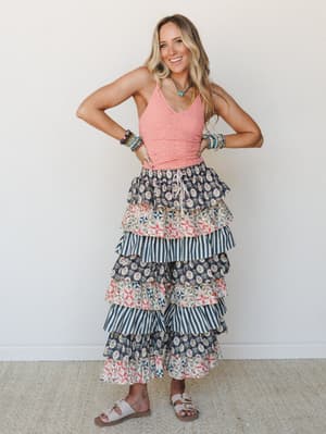 A smiling model in a coral crochet tank and show-stopping tiered ruffle pants — mixing floral block print, patchwork, and navy stripe tiers for a look that's equal parts festival energy and vintage quilt charm.