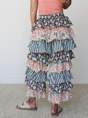 Shot from behind, a model wears statement tiered ruffle pants in a playful mix of floral, patchwork, and stripe prints layered from waist to ankle, paired with a coral textured tank and flat sandals — the kind of pants that do all the talking.