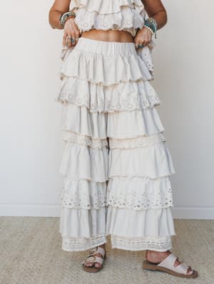 A tiered, ruffle-heavy wide-leg pant in soft taupe pairs with a matching eyelet crop top, layered beaded bracelets, and buckle sandals — the kind of outfit that feels like a daydream and still works for dinner.
