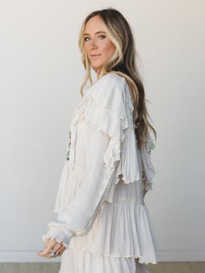 A cream boho blouse with cascading ruffle tiers and lace trim details, shot in soft studio light — the kind of top that feels equally at home at a bridal shower or a Sunday farmers market.