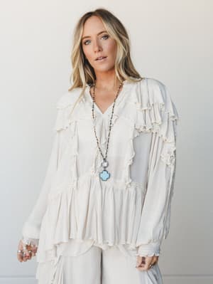 A cream ruffle blouse with layered petal tiers and lace trim worn loose and relaxed against a soft white studio wall — the kind of top that makes a simple outfit feel like it has a whole personality.