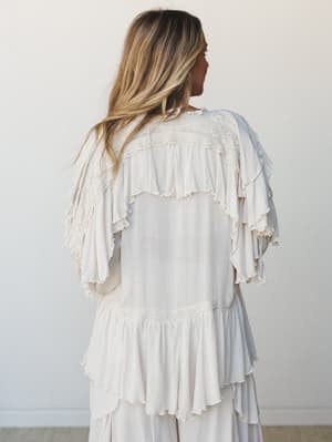 Shot from behind, a model in a cream ruffle-and-lace top with dramatic flutter sleeves stands against a soft white wall — the kind of piece that looks like it was made for slow mornings and something worth celebrating.