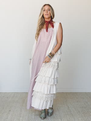 A layered boho look built around a cream tiered duster vest with eyelet and crochet trim worn over a dusty mauve printed maxi dress, styled with a red bandana, stacked bracelets, and teal western boots — free-spirited and effortlessly collected.