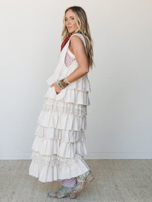 A tiered, eyelet-trimmed cream duster vest styled over a patterned bralette with cowboy boots and stacked bracelets — the kind of outfit that looks like it was pulled together effortlessly but absolutely wasn't.