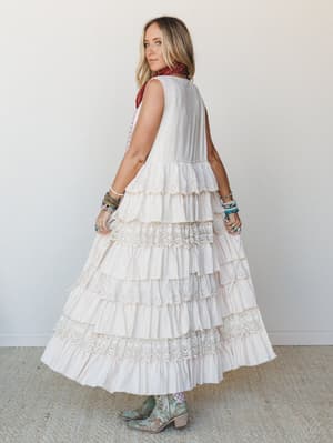 A cream tiered maxi duster vest with alternating eyelet and crochet lace trim layers, styled over a bandana and gingham with turquoise cowboy boots and stacked jewelry — the kind of outfit that looks like it took no effort but clearly took all the ri