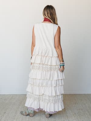 Shot from behind, a model wears a flowy tiered maxi duster vest in natural cream with eyelet lace trim between each ruffle layer, styled over a printed outfit with turquoise stacked bracelets, a red bandana neck scarf, and mint cowboy boots — effortl