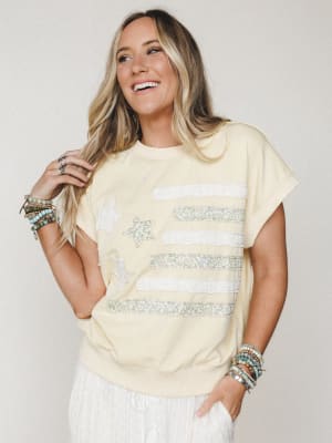 A soft butter yellow sweatshirt-style top with a patchwork flag graphic made from floral fabric strips and chenille star appliqués, styled with white wide-leg pants and stacked beaded bracelets — casual and expressive with a vintage-Americana feel.