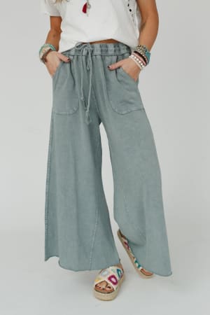 Wearing the Relaxing Robin Wide Leg Pant in Faded Teal with a white short-sleeve top, colorful bracelets, and multicolored woven sandals, this boho-comfy look is finished with a casual pose and hands in pockets.