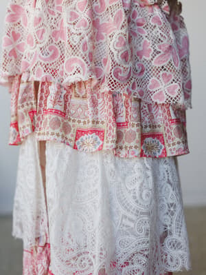 A dreamy close-up of layered lace and printed fabric tiers — pink embroidered crochet lace over a delicate floral block-print ruffle, falling into a cream paisley lace skirt layer, all soft texture and romantic detail.