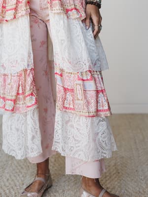 A close-up of a flowy tiered duster layered over blush pink pants, showing off the delicate lace overlay and block-print mixed panels — the kind of piece that makes a simple outfit feel like a whole moment.