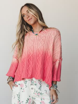A cable-knit sweater in a coral-to-pink ombre dip-dye styled with floral wide-leg pants and stacked beaded bracelets — the kind of outfit that looks effortless but clearly thought about.