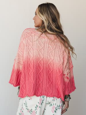 Shot from behind, a model in a cable-knit sweater with a soft coral-to-blush ombré fade pairs it with floral pants — the kind of effortless, pretty combination that feels like a good Saturday.
