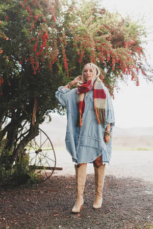 A woman stands outdoors under a flowering tree, wearing the Ophelia Oversized Denim Dress – Washed Denim, with tan knee-high boots and a red-and-white plaid scarf. An old wagon wheel behind her adds to the rustic, relaxed vibe.
