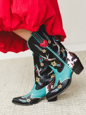 A close-up of statement cowboy boots in black and turquoise with tattoo-style embroidery — hearts, swallows, stars, and florals — paired with a billowy red skirt, shot against a soft neutral backdrop with serious personality.