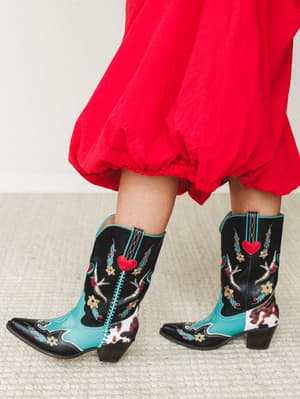 A close-up of bold embroidered cowboy boots in black, turquoise, and cowhide print, paired with a sweeping red midi skirt — the kind of combination that makes a room turn around.