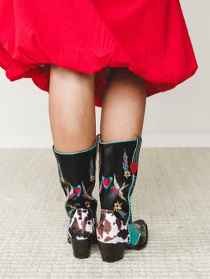 A back-view close-up of statement cowboy boots — black leather with bold embroidery, cowhide panels, turquoise piping, and heart and swallow motifs — paired with a billowing red skirt that hints at the full look.