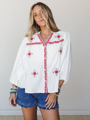 A white button-front blouse with colorful floral cross-stitch embroidery and pink geometric trim, styled with scallop-hem denim shorts and stacked boho bracelets — casual and expressive in the best way.