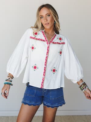 A white embroidered blouse with balloon sleeves and colorful floral cross-stitch detailing paired with scallop-hem denim shorts — the kind of top that does the work for you on a summer afternoon.