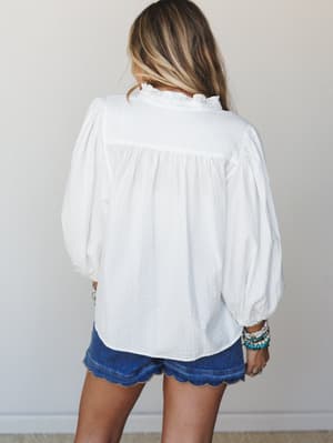 A breezy white blouse shot from the back, showing off its balloon sleeves and ruffle collar detail, paired with scallop-hem denim shorts and a stack of colorful beaded bracelets — effortless and a little romantic.