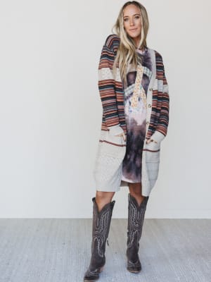 Woman wearing a long, patterned cardigan over a dress with cowboy boots on a plain background