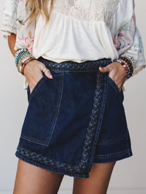 Dark denim wrap shorts with a standout braided trim detail along the waistband and hem, styled with a cream lace top, floral kimono, and stacked bracelets — the kind of outfit that looks effortless but clearly wasn't an accident.