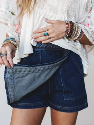A close-up mid-shot showing dark denim wrap-style shorts with a braided waistband detail, paired with a cream lace and floral top — the model's hands are stacked with beaded bracelets and silver rings, giving the whole thing that perfectly layered, c