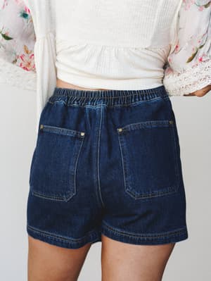 A back-view close-up of dark wash denim shorts with an elastic waistband and patch pockets, styled with a cropped white ribbed top and a floral kimono with lace trim — casual and put-together all at once.