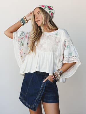 A breezy, layered boho look built around dark denim wrap shorts with braided trim, paired with a flowy mixed-fabric top featuring floral sleeves and lace paneling — finished with a crochet floral beanie and stacked bracelets for that effortlessly col