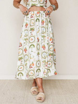 A flowy ivory midi skirt printed with framed tropical motifs — palm trees, pineapples, parrots, and botanicals — styled as a two-piece set with stacked rings, beaded bracelets, and raffia slide sandals for a warm-weather, free-spirited look.