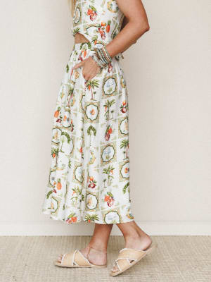 A cropped side view of a flowy ivory midi skirt covered in a whimsical tropical medallion print — palm trees, pineapples, and exotic fruit — styled as a matching set with raffia slide sandals and a stack of beaded bracelets.
