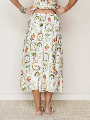 A breezy midi skirt in ivory with a vintage-inspired tropical print — palm trees, parrots, pineapples, and tropical fruit arranged in medallion frames — styled with a matching cropped top, stacked bracelets, and espadrilles, shot from the back agains