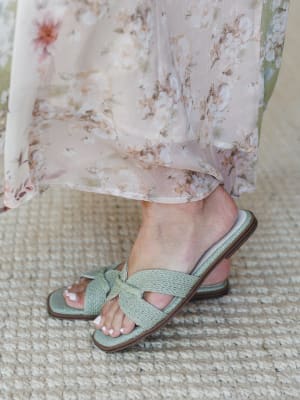 A close-up of sage green woven raffia slide sandals paired with a flowy floral maxi skirt in blush and cream — the kind of shoe-and-hem combo that makes you want to book a patio lunch immediately.