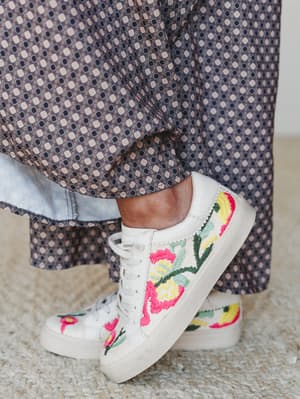A close-up of white lace-up sneakers covered in bold floral embroidery — hot pink, yellow, and sage green blooms — styled against the hem of a geometric-print wide-leg pant on a woven rug.
