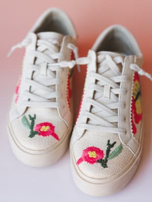 A pair of cream canvas lace-up sneakers covered in hand-embroidered floral details — pink blooms, yellow centers, green stems — shot up close against a soft blush backdrop with a dreamy, slightly romantic feel.