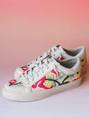 A pair of white low-top sneakers with vibrant floral embroidery across a woven textile panel, shot against a warm pink gradient background — the kind of shoes that make a sundress feel like a whole personality.