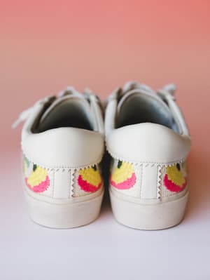 A pair of white low-top sneakers shot from the back heel angle, showing off colorful floral embroidery in hot pink, yellow, and green against a soft coral studio backdrop — the kind of shoes that do half the outfit work for you.
