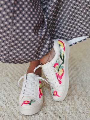 A close-up of white lace-up sneakers covered in bold floral embroidery — hot pink blooms, yellow centers, green leaves — worn with a geometric-print maxi skirt and standing on a woven jute rug.