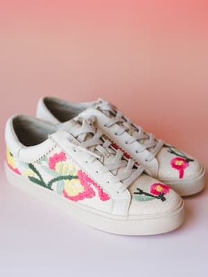 A pair of cream canvas sneakers covered in vibrant embroidered florals — hot pink blooms, sage leaves, and yellow accents — shot against a soft blush backdrop with the kind of light that makes you want to go somewhere pretty.