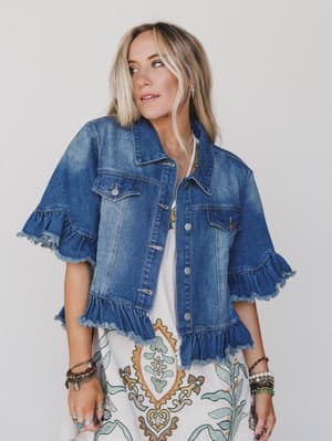 A woman wears the Scottie Ruffle Denim Jacket - Blue, featuring ruffled sleeves, flap pockets, and a cropped hem over a white patterned dress with layered bracelets, standing against a plain white background.