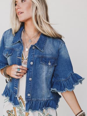 A woman wears the Scottie Ruffle Denim Jacket - Blue with ruffled sleeves, flap pockets, and a cropped hem, layered necklaces and rings over a white dress with a colorful floral print. Her long blonde hair flows as she looks to the side.