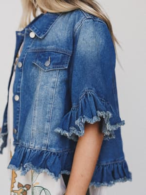 A woman wears the Scottie Ruffle Denim Jacket – Blue, featuring frayed ruffle trim on short sleeves and a cropped hem, paired with a patterned dress. Only her torso and part of her head are visible.