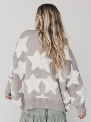 A person with long, wavy blonde hair is seen from behind, wearing the Wrapped In Stars Oversized Cardigan in taupe with large white stars and a green skirt, standing against a plain light background.