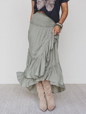 A person in a black T-shirt, layered bracelets, light tan cowboy boots, and the Washed Ashore Ruffled Maxi Skirt – Olive stands on a light-colored carpet for the perfect festival chic look.