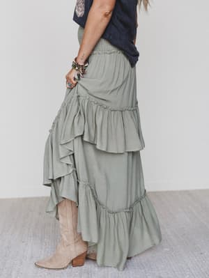A woman poses indoors in festival chic style, wearing the Washed Ashore Ruffled Maxi Skirt - Olive with a smocked waist, a dark top, bracelets, and light tan suede cowboy boots. She stands sideways, gently holding the ruffled skirt.