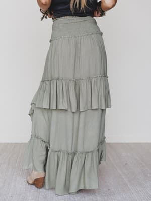 Someone stands indoors on a light floor, shown from the back, wearing the Washed Ashore Ruffled Maxi Skirt - Olive with a smocked waist and ruffle details, paired with brown shoes for a festival chic look.