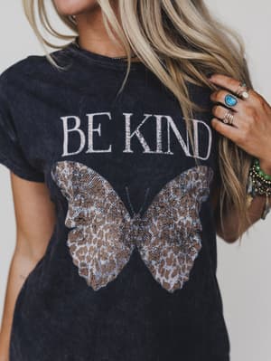 A person with long blonde hair wears the Be Kind Mineral Washed Graphic Tee in charcoal, featuring an oversized fit and a large butterfly graphic, styled with multiple rings and bracelets.