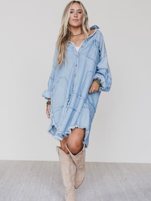 A woman with long blonde hair wears the Ophelia Oversized Denim Dress in washed denim, styled as a loose-fitting, light blue button-down and paired with tan knee-high boots against a plain white background.