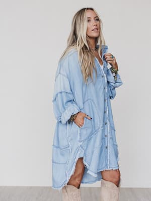 A woman with long blonde hair wears the Ophelia Oversized Denim Dress in washed denim, styled with tan knee-high boots and jewelry for a relaxed boho look. She stands sideways, one hand in her pocket, exuding effortless style.