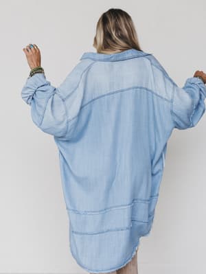 A person with long blonde hair faces away, wearing the Ophelia Oversized Denim Dress in washed denim with rolled-up sleeves. Arms raised, they display rings and bracelets against a plain white background.
